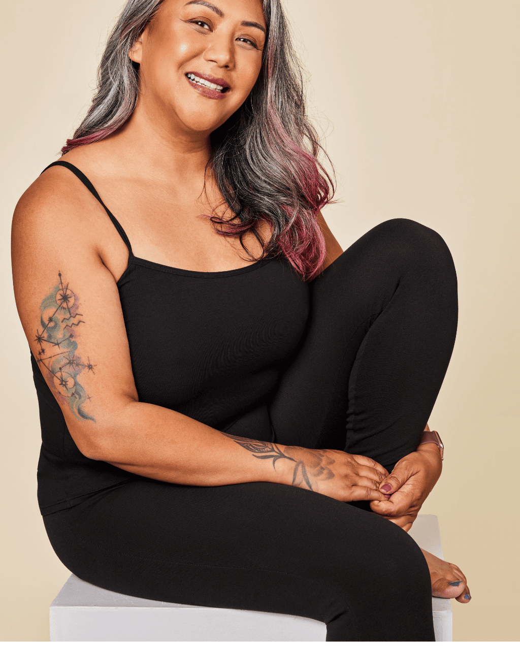 Woman wearing a black sleeveless top and leggings sitting on a white stool against a white background