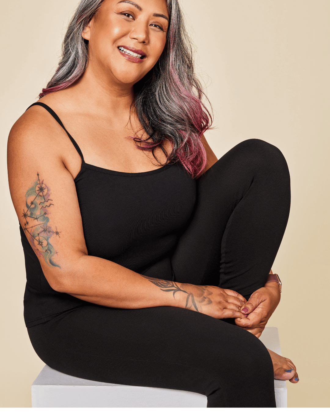 Woman wearing a black sleeveless top and leggings sitting on a white stool against a white background
